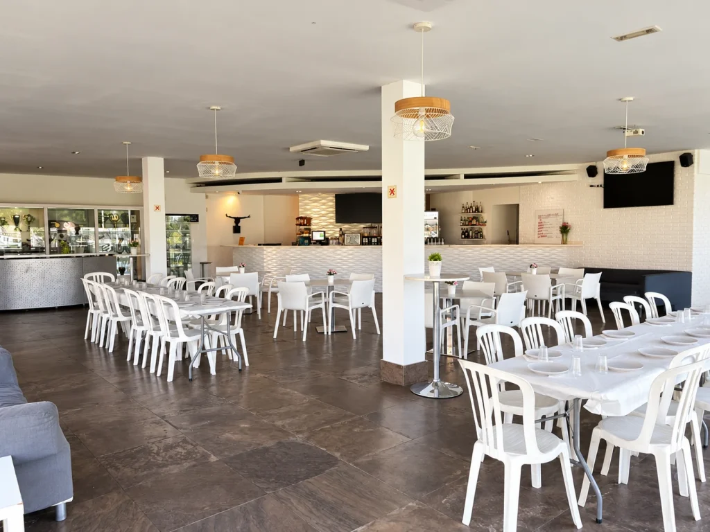 Sala de la cafetería de La Colina con mesas, sillas blancas y barra al fondo.